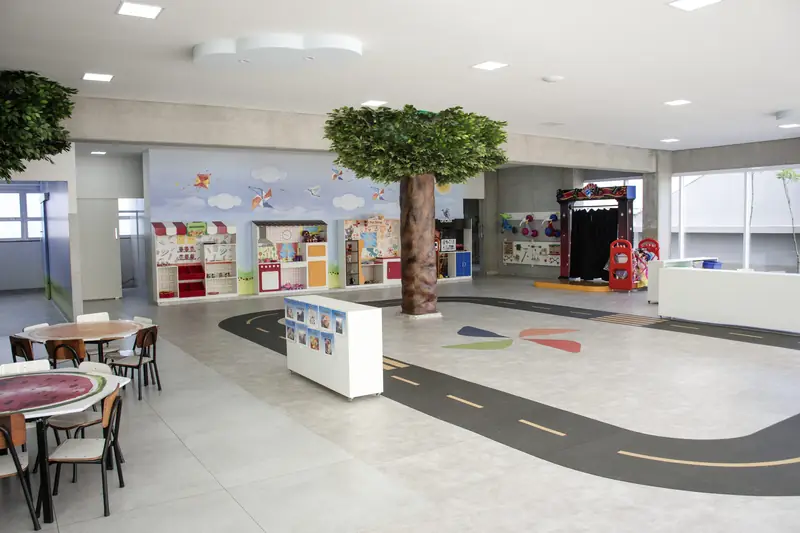 Espaço amplo e iluminado da Educação Infantil, com árvore cenográfica e mesas baixas