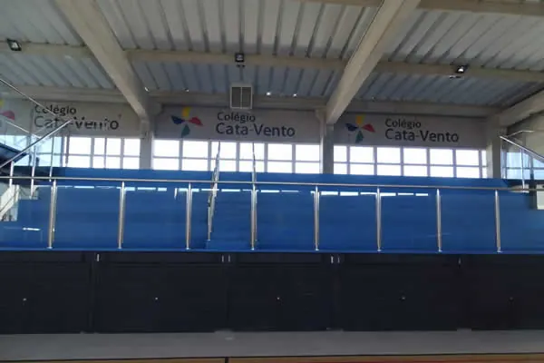 Quadra coberta com arquibancada azul e logo do colégio na parede