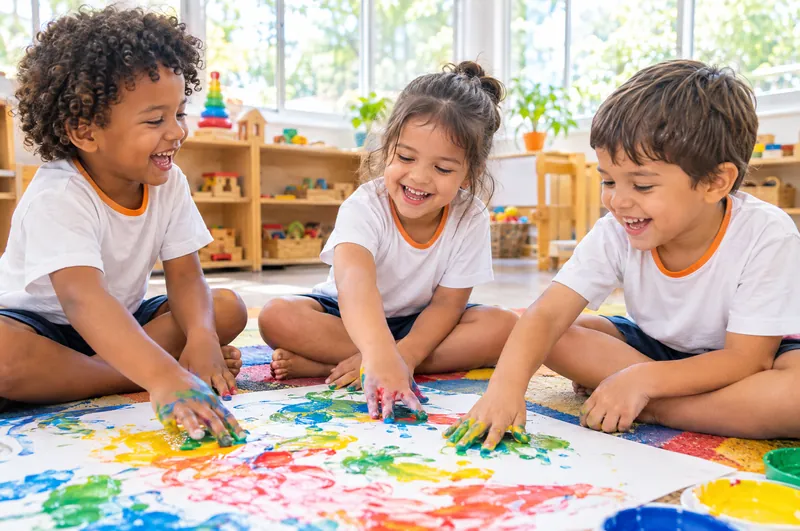 Três crianças da Educação Infantil pintam com as mãos sobre um papel colorido, sentadas no tapete da sala